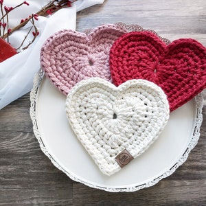 May include: Three crocheted heart-shaped coasters in pink, red, and white on a white tray with a lace trim. The coasters are handmade and have a textured surface.