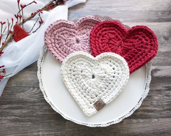 May include: Three crocheted heart-shaped coasters in pink, red, and white on a white tray with a lace trim. The coasters are handmade and have a textured surface.