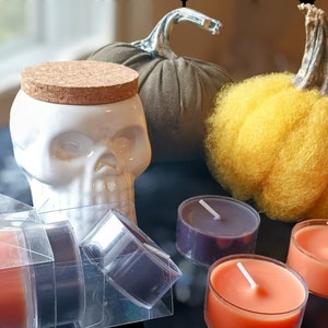 Op de afbeelding: Een witte keramische schedelvormige container met een kurken deksel, omringd door decoratieve pompoenen en theelichtkaarsen in oranje en paars. De verpakking bevat de tekst "Spooky time!"