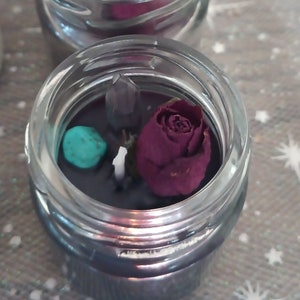 May include: Small glass jar candles with a dark wax base, each containing a dried burgundy rose, a small turquoise stone, and a clear crystal. The candles are arranged in a row. The background is soft focus.