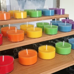 Op de afbeelding: Een verzameling kleurrijke waxinelichtjes gerangschikt op een bamboe display met meerdere niveaus. De kaarsen zijn in de kleuren rood, oranje, geel, groen, blauw en paars. Elke kaars heeft een witte lont.