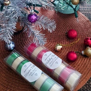 Könnte beinhalten: Zwei Sets Duftkerzen in durchsichtiger Plastikverpackung. Die Kerzen sind in einem Stapel von vier angeordnet. Die Kerzen sind mit "Winter Vibe" und "Holiday Tree" beschriftet. Die Etiketten enthalten auch die Duftbeschreibungen: "Gefallene Blätter, Balsamtanne" und "Weihnachtskuchen, Gewürzter Glühwein".