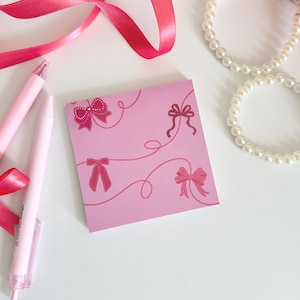 May include: Pink notepad with a pink ribbon and a pink bow design. The notepad has a white background with pink lines and pink bows. The notepad is on a white surface.