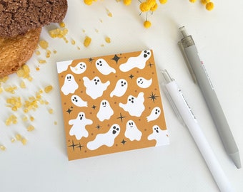 May include: A square notepad with a repeating pattern of white cartoon ghosts on an orange background. Two pens, one white and one gray, are next to the notepad. Two cookies and yellow flowers are also in the image.