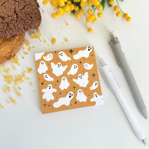 May include: A patterned notepad featuring white ghost illustrations on an orange backdrop. Two pens, one white and one grey, are positioned beside the notepad. Two biscuits and yellow flowers are also visible in the image.