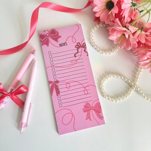 May include: Pink notepad with a checklist and pink bows. The notepad has the word "NOTES" printed on it. The notepad is on a white surface with pink ribbon and pearl necklaces.