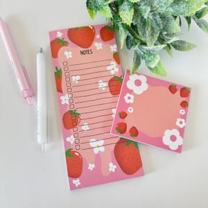 May include: Two pink notepads with strawberry and flower designs. The larger notepad has a section for notes and a checklist. The smaller notepad is blank.