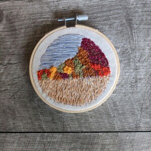 May include: Embroidered hoop art featuring a landscape scene with a blue sky, brown hills, and colorful trees. The trees are embroidered in shades of red, orange, yellow, and green.