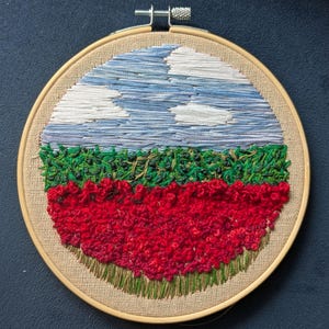 May include: A circular embroidery hoop with a landscape scene. The top of the hoop features a blue sky with white clouds. The middle of the hoop features a green field with a row of red flowers. The bottom of the hoop features a row of green grass.