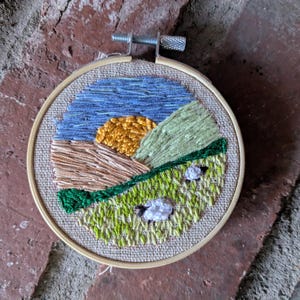 May include: Embroidered hoop art depicting a landscape with a blue sky, yellow sun, green hills, and white sheep.