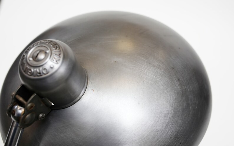 May include: A silver metal desk lamp with a round shade and a knob with the text "Original Design" on it.