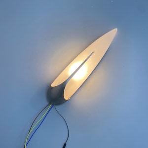 Peut inclure: Une applique murale moderne au design unique. Le luminaire a une forme incurvée et allongée avec une source de lumière centrale. L'applique est fixée sur une base grise et comporte des fils apparents. La lumière émet une lueur chaude sur un mur bleu.