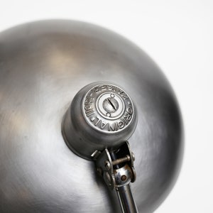 May include: A silver metal desk lamp with a round shade and a swivel arm. The lamp has a metal base and a metal shade. The shade has a small hole in the centre. The lamp is labelled "ORIGINAL ALL METAL REPRODUCED".