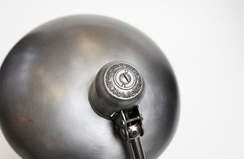 May include: A silver metal desk lamp with a round shade and a swivel arm. The lamp has a metal base and a metal shade. The shade has a small hole in the centre. The lamp is labelled "ORIGINAL ALL METAL REPRODUCED".