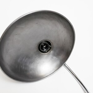 May include: A silver metal desk lamp with a large, round, reflective shade. The lamp is switched on and the light bulb is visible.