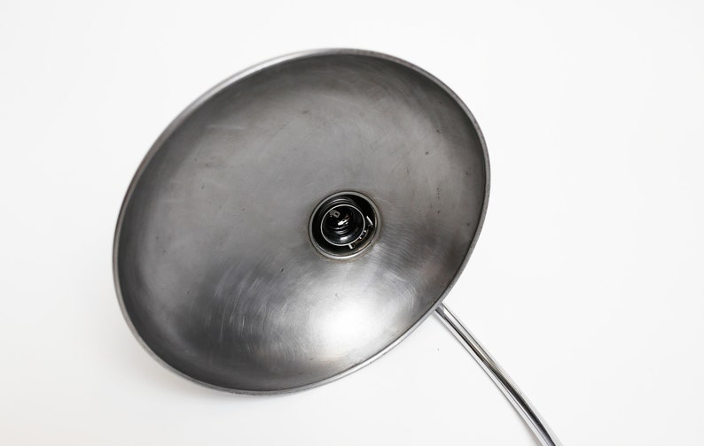 May include: A silver metal desk lamp with a large, round, reflective shade. The lamp is switched on and the light bulb is visible.