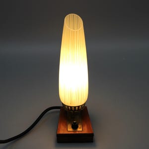 Könnte beinhalten: Eine Vintage-Tischlampe mit geripptem, cremefarbenem Glasschirm. Die Lampe steht auf einem Holzsockel mit Messingakzenten und einem schwarzen Netzkabel. Die Lampe strahlt ein warmes Licht aus, geeignet für einen Nachttisch oder Schreibtisch.