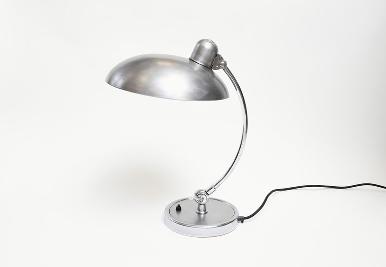 May include: A silver desk lamp with a curved arm and a round shade. The lamp has a black cord and a switch on the base.