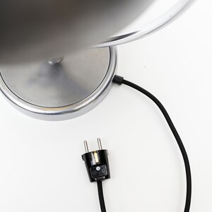 May include: A silver table lamp with a round base and a black cord with a plug. The lamp has a silver shade.