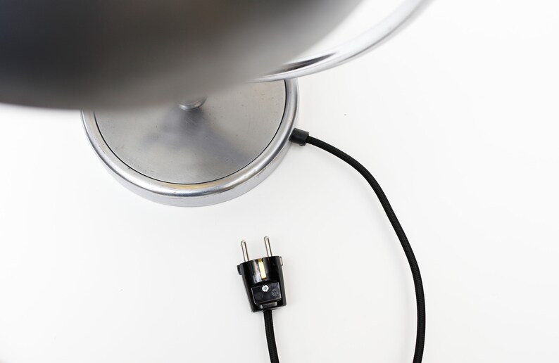 May include: A silver table lamp with a round base and a black cord with a plug. The lamp has a silver shade.