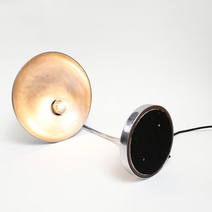 May include: A silver desk lamp with a round, metallic shade and a black base. The lamp is switched on and the light is shining through the shade.