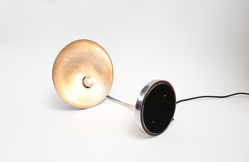 May include: A silver desk lamp with a round, metallic shade and a black base. The lamp is switched on and the light is shining through the shade.