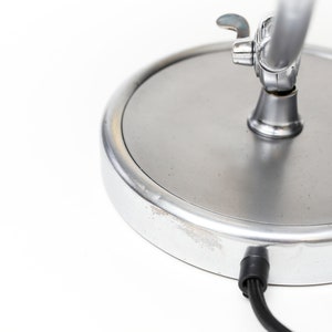 May include: A silver desk lamp with a round base and a flexible arm. The lamp has a switch on the arm and a cord coming out of the base.