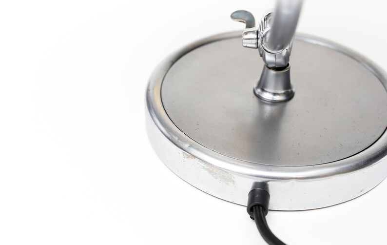 May include: A silver desk lamp with a round base and a flexible arm. The lamp has a switch on the arm and a cord coming out of the base.