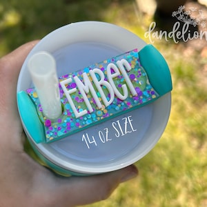 May include: A turquoise and white 14 oz tumbler with a clear straw and a turquoise and glitter name tag that says "EMBER".