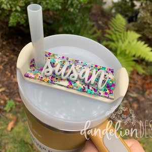May include: A white and gold tumbler with a clear lid and a straw. The lid has a clear, rectangular name tag with the name "Susan" written in white letters and a colorful confetti design. The tumbler has a black handle and the words "dandelion DESIGN" are printed on the side.