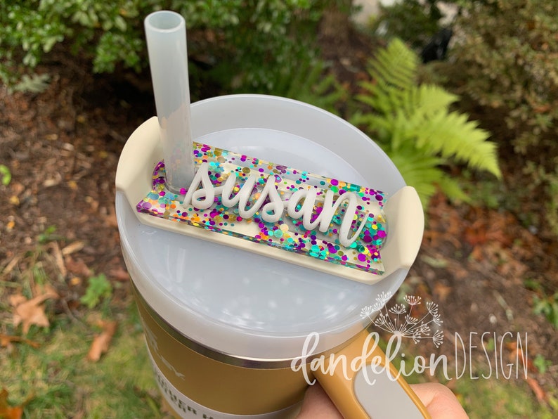 May include: A white and gold tumbler with a clear lid and a straw. The lid has a clear, rectangular name tag with the name "Susan" written in white letters and a colorful confetti design. The tumbler has a black handle and the words "dandelion DESIGN" are printed on the side.