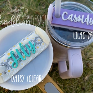 May include: Two personalized straw covers for tumblers. One is clear with a daisy pattern and the name "Lilly" in teal. The other is lilac pastel with the name "Cassidy" in white.