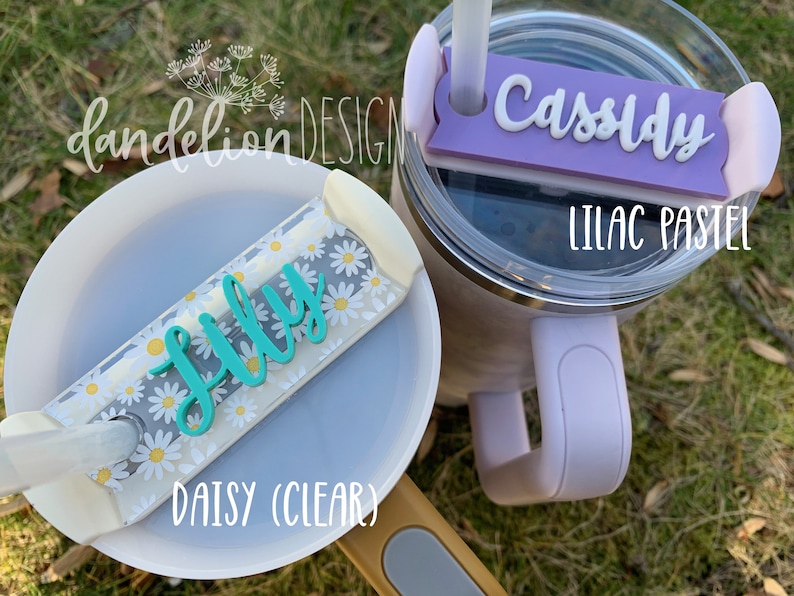 May include: Two personalized straw covers for tumblers. One is clear with a daisy pattern and the name "Lilly" in teal. The other is lilac pastel with the name "Cassidy" in white.