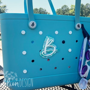May include: A turquoise tote bag with white polka dots and a custom letter "B" design. The bag has two handles and a pair of purple and white swim goggles hanging from the side. The bag is near a pool.