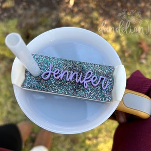 May include: A white tumbler with a clear lid and a straw. The tumbler has a glitter-covered name tag that says "Jennifer" in purple lettering.