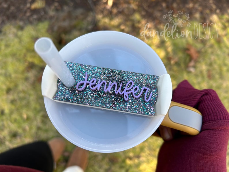 May include: A white tumbler with a clear lid and a straw. The tumbler has a glitter-covered name tag that says "Jennifer" in purple lettering.