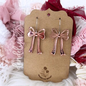 May include: A pair of rose gold bow earrings on a brown card with the text "dandelion design".