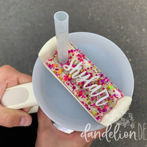 May include: A white plastic cup with a clear lid and a straw. The lid has a pink, yellow, and gold glitter design with the name "Skyla" written in white letters.