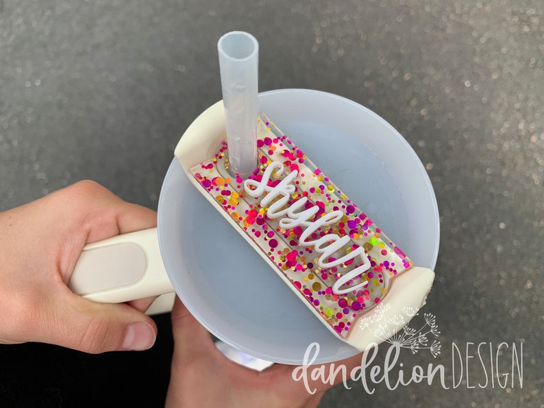 May include: A white plastic cup with a clear lid and a straw. The lid has a pink, yellow, and gold glitter design with the name "Skyla" written in white letters.