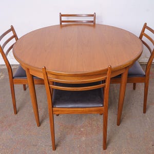 May include: A round wooden dining table with a smooth, light brown grain finish, accompanied by four matching wooden chairs. Each chair features a curved backrest with two horizontal slats and a black upholstered seat cushion for support.