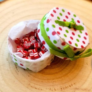 May include: A small round paper box with a white lid featuring red and green polka dots, tied with a green ribbon. The box is lined with white tissue paper and filled with small, cylindrical red and white striped candies.
