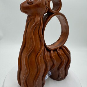 Könnte beinhalten: Eine braune Keramikskulptur eines Lamas mit einem großen kreisförmigen Griff. Das Lama hat einen langen Hals und Körper mit wellenförmigen Linien und einen kleinen Kopf mit detaillierten Merkmalen. Die Skulptur steht auf einer weißen Oberfläche.