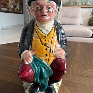 May include: A ceramic figurine of an elderly man wearing a black hat, glasses, and a yellow vest. He holds two red sacks, one covered with a green cloth. The figurine has a detailed, hand-painted design.