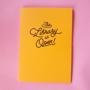 Puede incluir: Un cuaderno amarillo brillante con la frase "The Library is Open!" en escritura negra. El cuaderno est&aacute; sobre un fondo rosa, creando un contraste. El texto de la portada est&aacute; en un estilo manuscrito juguet&oacute;n.