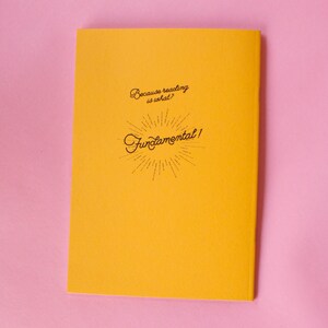 Puede incluir: Un cuaderno amarillo con el texto "Because reading is what? Fundamental!" en escritura negra. El cuaderno est&aacute; sobre una superficie rosa.