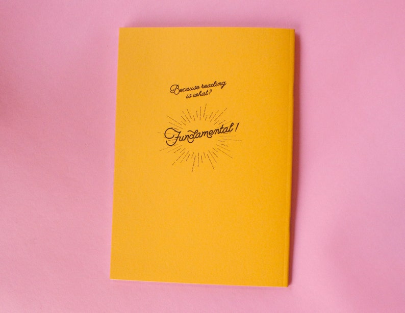 Puede incluir: Un cuaderno amarillo con el texto "Because reading is what? Fundamental!" en escritura negra. El cuaderno est&aacute; sobre una superficie rosa.