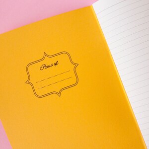 Puede incluir: Cuaderno abierto con una cubierta amarilla brillante y p&aacute;ginas rayadas. La cubierta presenta una etiqueta decorativa con el texto "Haus of" y espacio para un nombre. Fondo rosa.