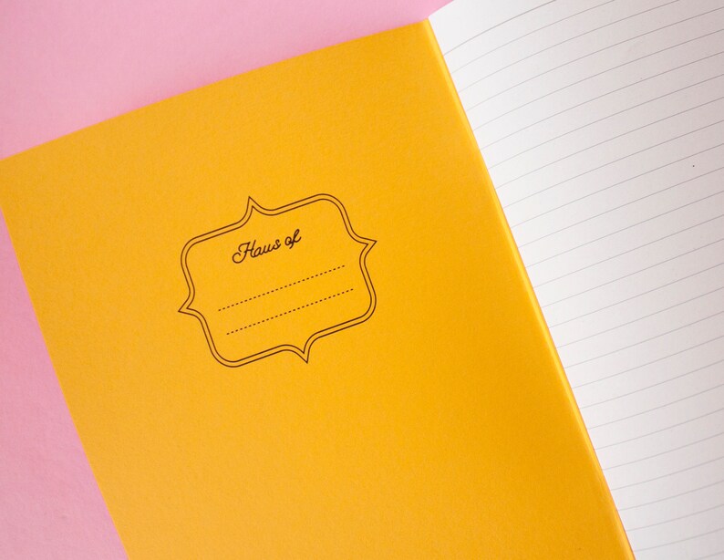 Puede incluir: Cuaderno abierto con una cubierta amarilla brillante y p&aacute;ginas rayadas. La cubierta presenta una etiqueta decorativa con el texto "Haus of" y espacio para un nombre. Fondo rosa.