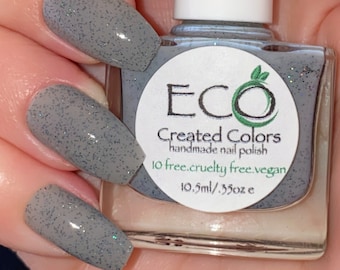 May include: A hand model wearing grey glitter nail polish. The bottle of nail polish is labelled "ECO Created Colors handmade nail polish 10 free. cruelty free. vegan. 10.5ml / .35oz e"