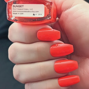 May include: A bottle of bright orange nail polish labeled "SUNSET" by Eco Created Colors, LLC. The bottle is in a hand with four orange nails painted with the polish.